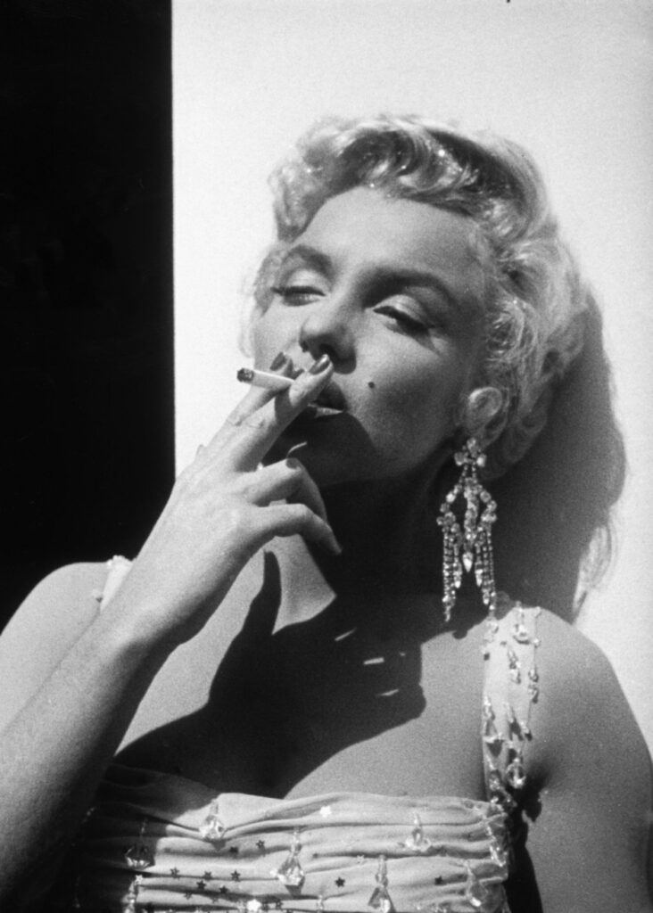 Black and white photo of Marilyn Monroe smoking a cigarette and wearing long gem studded earrings.